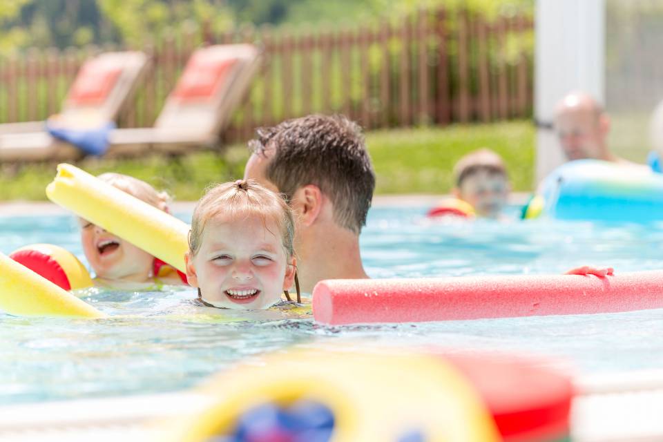 Children with parents in the pool with swimming noodles.