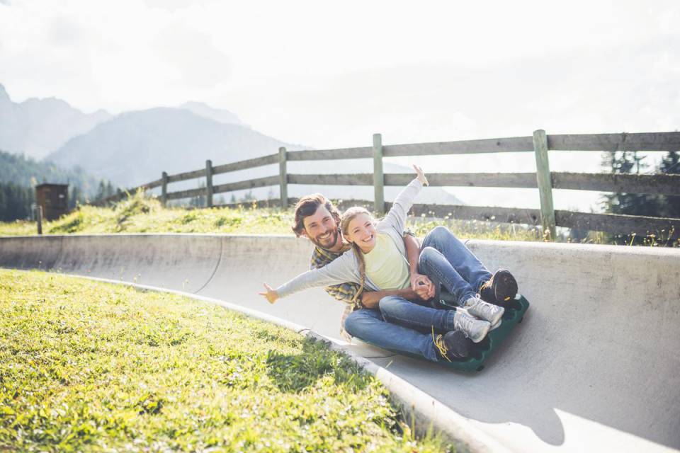 Ein lachender Vater fährt mit seiner Tochter auf einer Sommerrodelbahn hinunter. 