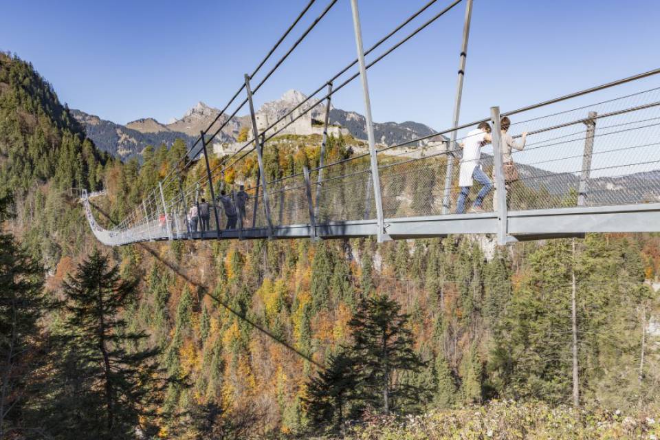 Hängebrücke über bewaldetes Tal mit Herbstfarben, zwei Personen laufen darauf, Berge im Hintergrund