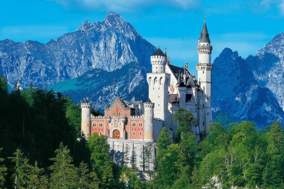 Schloss Neuschwanstein vor Alpenkulisse und blauem Himmel, umgeben von grünen Wäldern