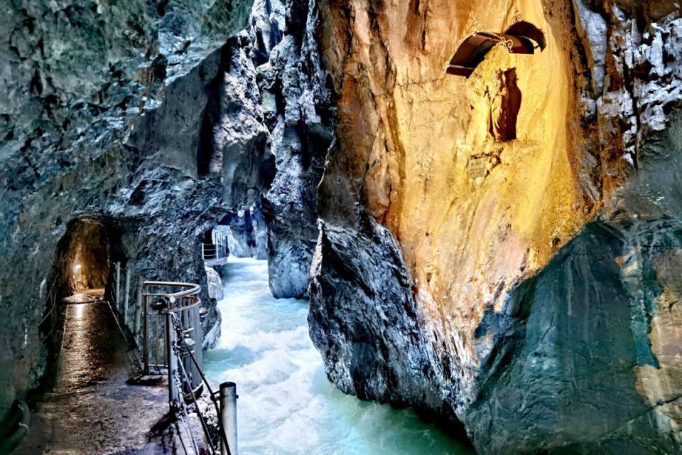 Schmaler Wandersteig mit Geländer entlang tosender, türkisfarbener Wildwasserströmung in einer hohen Felsklamm – begehbarer Steg in natürlicher Höhle.