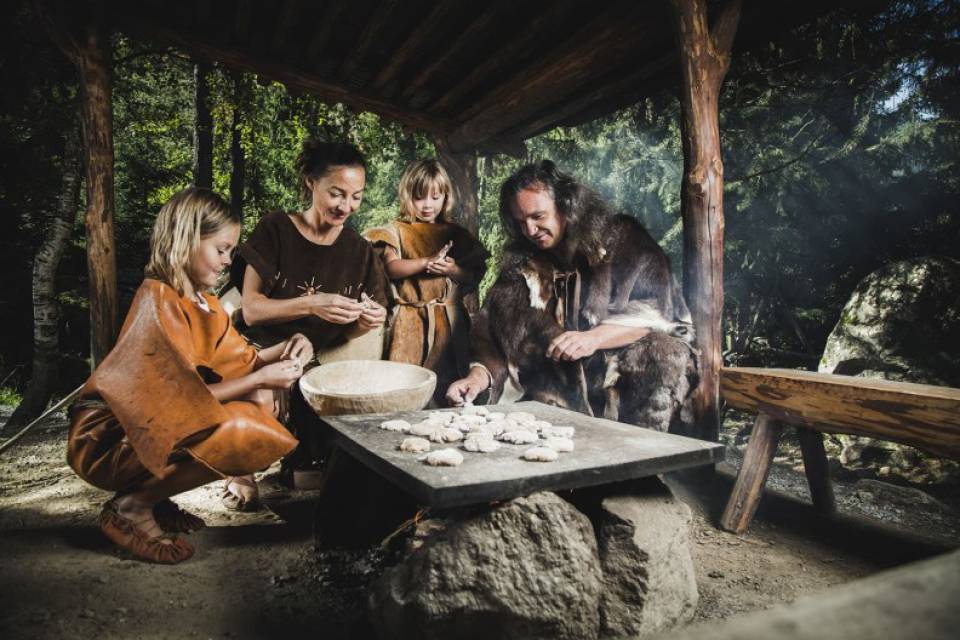 Besucher in steinzeitlicher Kleidung im Ötzi-Dorf Freilichtmuseum: zwei Kinder in einfachen Ledergewändern und zwei Erwachsene mit Fellumhängen formen Teigfladen auf einer großen Steinplatte unter einem hölzernen Unterstand vor dichter Waldkulisse