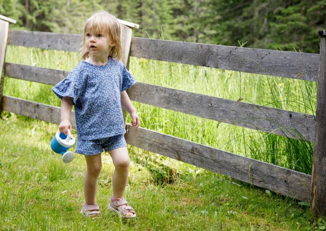 Kleines Mädchen läuft mit Gießkanne auf Wiese an einem Zaun entlang