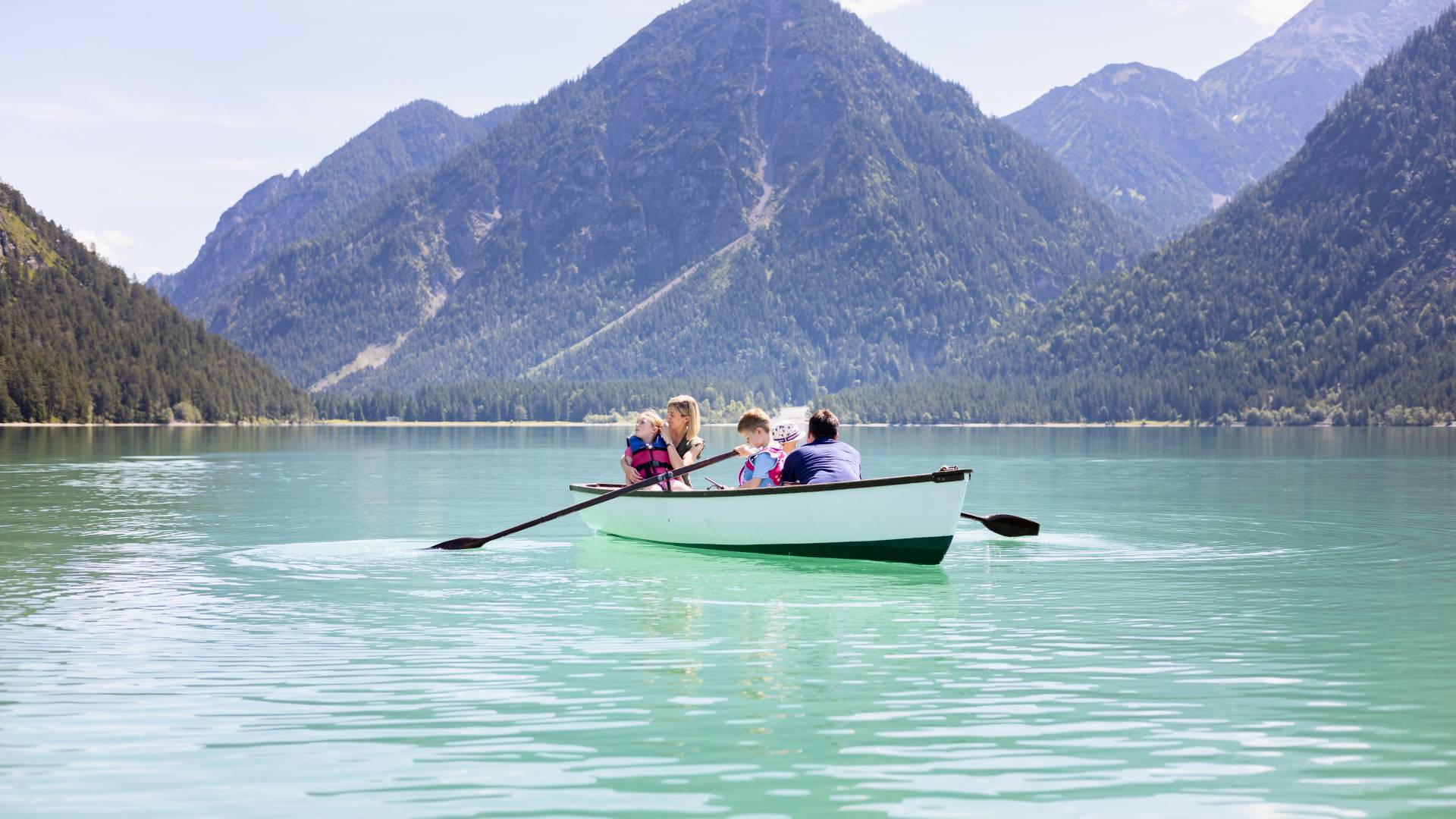 Familie sitzt im Boot auf klarem See mit Blick auf die Tiroler Alpen