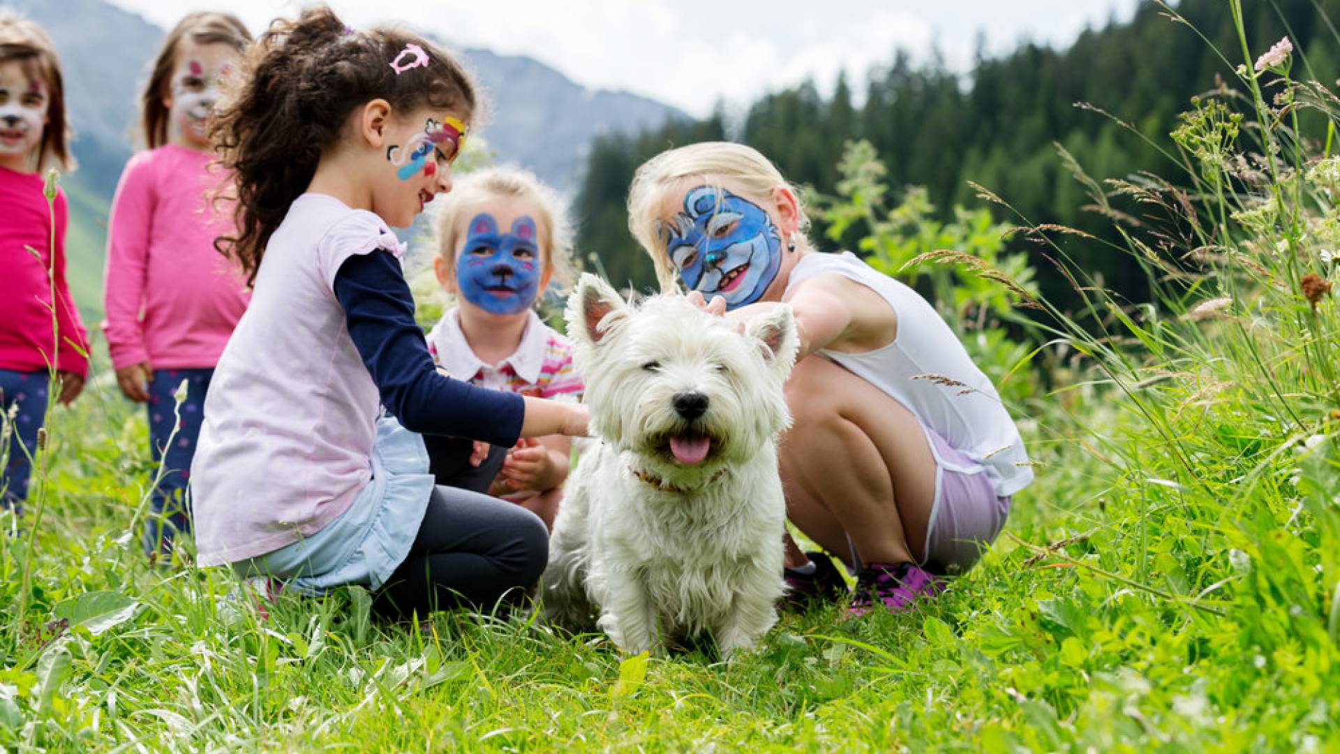 Fröhliche, bunt geschminkte Kinder streicheln einen kleinen weißen Hund auf einen Wiese vom Hotel Kaiserhof